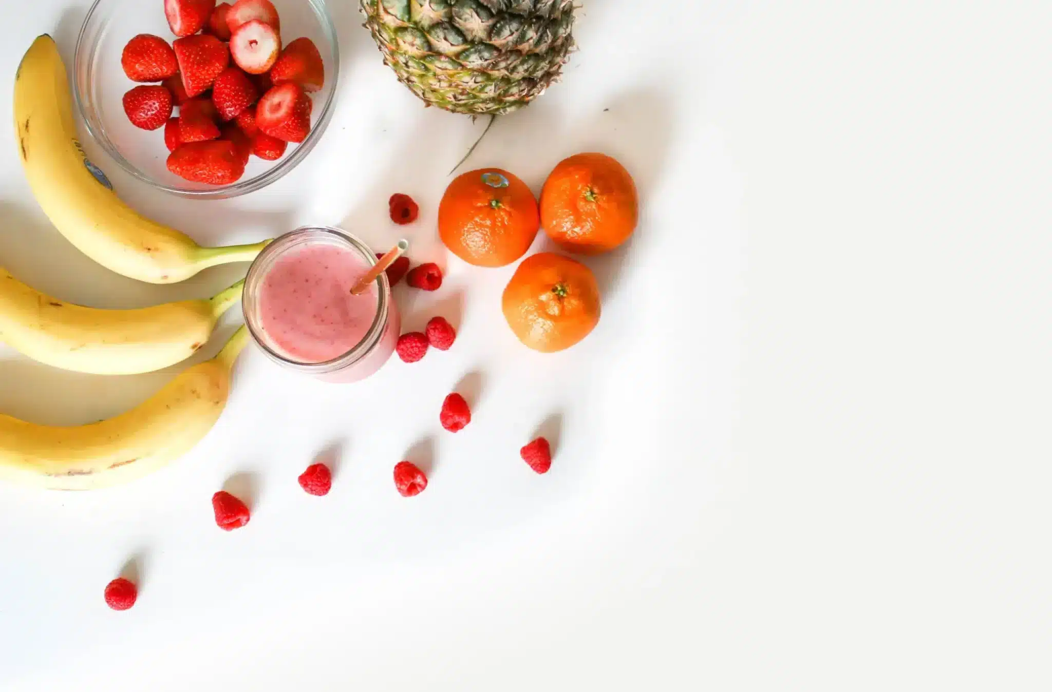 Um arranjo brilhante e saudável de frutas numa superfície branca, com bananas maduras, um ananás, várias tangerinas, uma taça de morangos, framboesas espalhadas e um copo de batido cor-de-rosa com uma palhinha.
